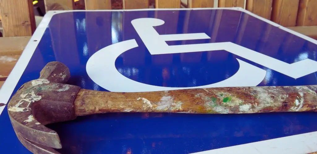 Obrazek do artykułu - co daje lekki stopień niepełnosprawności. Na zdjęciu widoczny jest znak informujący o miejscu dla osób niepełnosprawnych, przedstawiony na niebieskim tle z białym symbolem wózka inwalidzkiego. Na znaku leży zużyty młotek z drewnianym, poplamionym farbą trzonkiem i metalową głowicą. Tło zdjęcia stanowi drewniana konstrukcja, która wydaje się być elementem ogrodzenia lub budynku. Połączenie tych elementów nadaje zdjęciu symboliczny charakter, mogący wskazywać na temat związany z naprawą, dostępnością lub prawami osób niepełnosprawnych.