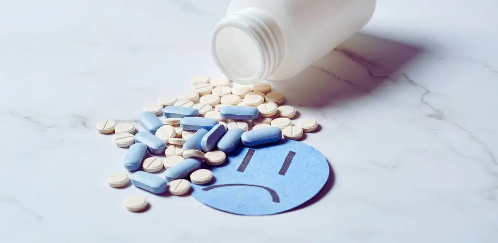 obrazek do artykułu - czy depresja jest uleczalna. Obraz przedstawia rozsypane tabletki – zarówno białe, jak i niebieskie – wychodzące z przewróconej białej butelki na tle białej, marmurowej powierzchni. Wśród tabletek leży niebieskie kółko z narysowaną smutną miną, symbolizujące przygnębienie lub depresję.