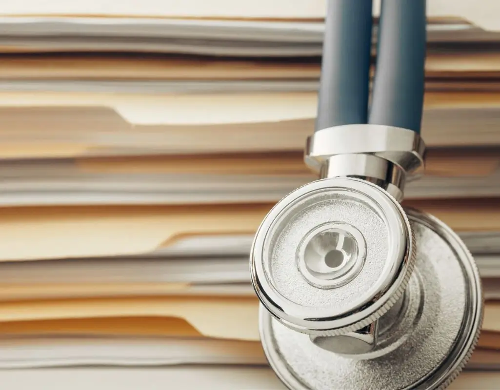 Obrazek do artykułu - orzeczenie o niepełnosprawności. Na zdjęciu widać stos dokumentów, które są ułożone w schludny stos, a na nich oparty jest stetoskop. Obraz przedstawia połączenie tematyki medycznej z formalnymi dokumentami, co sugeruje związki między zdrowiem a dokumentacją prawną lub administracyjną, taką jak orzeczenia lekarskie czy dokumentacja związana z opieką zdrowotną. Stetoskop z metalową głowicą i niebieskim przewodem dodaje element medyczny, co może nawiązywać do formalnych procedur oceny zdrowia lub diagnostyki w kontekście prawnym.