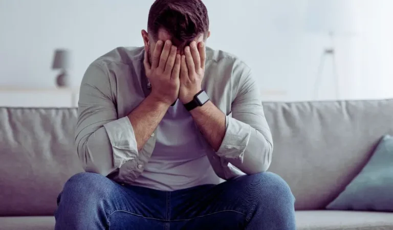 Mężczyzna siedzący na kanapie z twarzą schowaną w dłoniach, wyrażający emocje takie jak smutek i frustracja. Idealne zdjęcie do treści o tematyce praca dla chorych psychicznie, ukazujące trudności i wyzwania psychiczne.