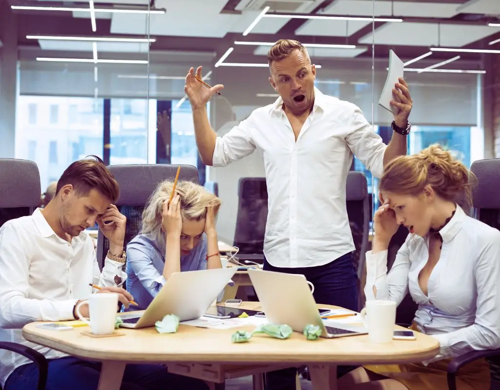 Na obrazie widać cztery osoby siedzące przy stole w biurze. Trzy z nich wyglądają na zestresowane, zmęczone lub przytłoczone. Mają pochylone głowy, trzymają się za czoło i sprawiają wrażenie, jakby intensywnie myślały lub przeżywały trudną sytuację. Przed nimi na stole leżą laptopy, kubki i pogniecione kartki, co może sugerować, że są w trakcie burzliwego spotkania lub projektu, który nie idzie zgodnie z planem. Za nimi stoi mężczyzna, który gestykuluje i wygląda na wzburzonego lub zdenerwowanego – możliwe, że podnosi głos. Jego postawa i wyraz twarzy sugerują frustrację, co może potęgować napięcie w grupie. Atmosfera na obrazie jest pełna stresu, konfliktu i napięcia, co może oddawać trudne relacje, mobbing w pracy lub presję zawodową.