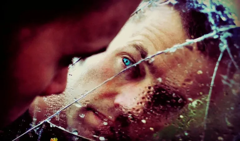 Zdjęcie przedstawia osobę wpatrującą się w swoje odbicie w popękanym lustrze. Rysy twarzy są zniekształcone przez linie pęknięć i krople wody, co nadaje obrazowi surrealistyczny, niemal malarski charakter. Intensywnie błękitne oko wyróżnia się na tle przytłumionych barw, przyciągając uwagę i budząc skojarzenia z introspekcją oraz poszukiwaniem prawdy o sobie. Popękane lustro może symbolizować kruchość ludzkiego umysłu, moment wewnętrznego rozdarcia lub spojrzenie na świat przez pryzmat innej wrażliwości. To subtelne napięcie, które wywołuje choroba psychiczna i kreatywność jest tu niemal namacalne – obraz balansuje na granicy niepokoju i artystycznej głębi, ukazując, jak emocje i percepcja mogą kształtować sposób postrzegania rzeczywistości.