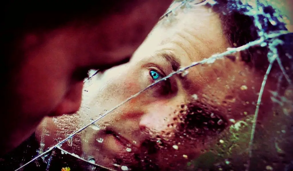 Zdjęcie przedstawia osobę wpatrującą się w swoje odbicie w popękanym lustrze. Rysy twarzy są zniekształcone przez linie pęknięć i krople wody, co nadaje obrazowi surrealistyczny, niemal malarski charakter. Intensywnie błękitne oko wyróżnia się na tle przytłumionych barw, przyciągając uwagę i budząc skojarzenia z introspekcją oraz poszukiwaniem prawdy o sobie. Popękane lustro może symbolizować kruchość ludzkiego umysłu, moment wewnętrznego rozdarcia lub spojrzenie na świat przez pryzmat innej wrażliwości. To subtelne napięcie, które wywołuje choroba psychiczna i kreatywność jest tu niemal namacalne – obraz balansuje na granicy niepokoju i artystycznej głębi, ukazując, jak emocje i percepcja mogą kształtować sposób postrzegania rzeczywistości.