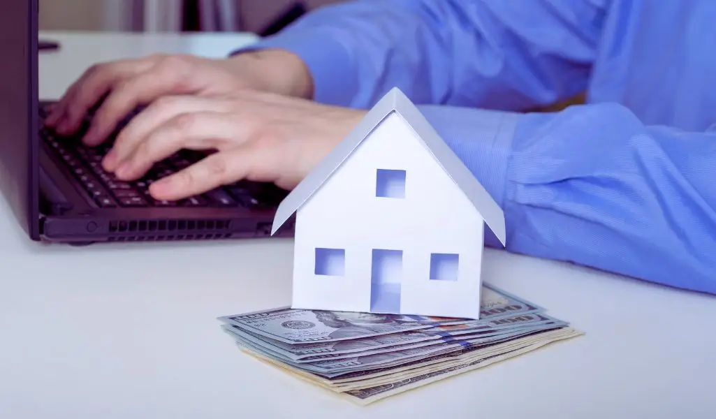 Na zdjęciu widać osobę ubraną w niebieską koszulę, piszącą na laptopie. Na pierwszym planie leży sterta banknotów, na której stoi symboliczny biały domek wykonany z papieru. Obrazek budzi skojarzenia z tematyką finansowania mieszkań, planowania budżetu i poszukiwania wsparcia finansowego. Może symbolizować bezpieczeństwo, marzenia o własnym lokum oraz proces ubiegania się o pomoc finansową. Idealnie oddaje temat dofinansowanie do wynajmu mieszkania dla niepełnosprawnych, wskazując na formalności, wsparcie i realne środki potrzebne do zapewnienia stabilnego miejsca do życia.