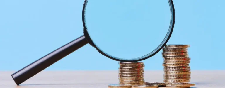 Na obrazie widać stosy ułożonych monet w złotym kolorze, które symbolizują finanse i oszczędności. Przed nimi znajduje się powiększające szkło, które skupia uwagę na szczegółach i sugeruje analizę lub badanie finansowe. Tło jest jasnoniebieskie, co nadaje całości wrażenie profesjonalizmu i przejrzystości. Obraz oddaje uczucie staranności, dokładności oraz nadziei na lepsze zarządzanie środkami. Idealnie ilustruje temat "Dofinansowanie wynagrodzeń PFRON w 2025", sugerując skrupulatne przyglądanie się możliwościom wsparcia finansowego oraz planowaniu w przyszłości.