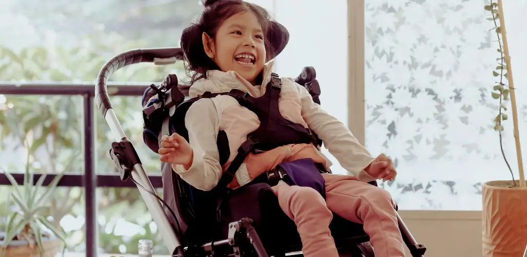 Na zdjęciu widać uśmiechniętą dziewczynkę siedzącą w specjalistycznym wózku, który zapewnia jej stabilizację i wsparcie. Jej radość jest zaraźliwa – szeroki uśmiech i błyszczące oczy pokazują beztroskę chwili. Wózek sugeruje, że dziecko dotknęła niepełnosprawność sprzężona, jednak nie odbiera jej energii i dziecięcej radości. Jasne, pełne światła tło z roślinami tworzy ciepłą, przyjazną atmosferę, podkreślającą pozytywne emocje.