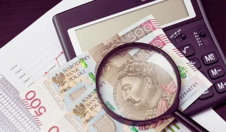 Na zdjęciu widzimy kilka banknotów o nominale 500 zł z wizerunkiem króla Jana III Sobieskiego, częściowo powiększonych przez szkło powiększające. Banknoty leżą na dokumentach z tabelami i wykresami finansowymi, a obok znajduje się kalkulator. Całość tworzy atmosferę analizy finansowej, planowania budżetu lub zastanawiania się nad wynagrodzeniem. Szkło powiększające symbolizuje dokładne przyglądanie się finansom, co może odnosić się do tematu "ile zarabia osoba z orzeczeniem" i analizy dochodów osób z niepełnosprawnościami. Zdjęcie wywołuje skojarzenia z liczeniem, budżetowaniem i rozważaniem różnych źródeł dochodu.