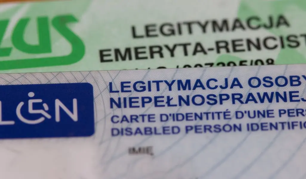 Zdjęcie przedstawia dwie plastikowe karty tożsamości, leżące jedna na drugiej. Na pierwszym planie widoczna jest legitymacja osoby niepełnosprawnej, oznaczona niebieskim symbolem osoby na wózku inwalidzkim. Napisy na karcie są wyraźne, zarówno w języku polskim, jak i francuskim oraz angielskim. W tle częściowo widoczna jest legitymacja emeryta-rencisty, wydana przez ZUS, w odcieniach zieleni. Obie karty są lekko nachylone, co dodaje dynamiki obrazowi. Zdjęcie oddaje powagę i ważność tych dokumentów – stanowią one symbol przynależności do grupy osób wymagających specjalnego wsparcia i uprawnień. Widać w nich zarówno aspekt formalny, jak i pewne emocjonalne znaczenie – towarzyszą one codziennemu życiu ich posiadaczy, niosąc zarówno ulgę w trudach, jak i przypomnienie o istniejących barierach.