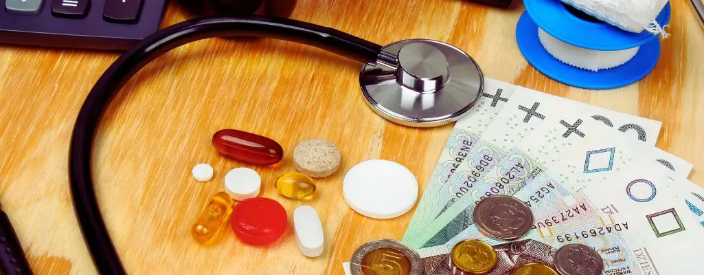 Na drewnianym stole leży stetoskop, różnorodne tabletki i kapsułki, a obok nich banknoty i monety polskiej waluty. W tle widać rolkę bandaża oraz kalkulator, co sugeruje kontekst finansowy związany z opieką zdrowotną. Obrazek symbolizuje wysokie koszty leczenia i potrzebę wsparcia finansowego, zwracając uwagę na problematykę zwrot za leki dla niepełnosprawnych. Kolory są ciepłe, ale zestawienie pieniędzy i leków budzi refleksję nad dostępnością terapii i finansowymi trudnościami pacjentów.