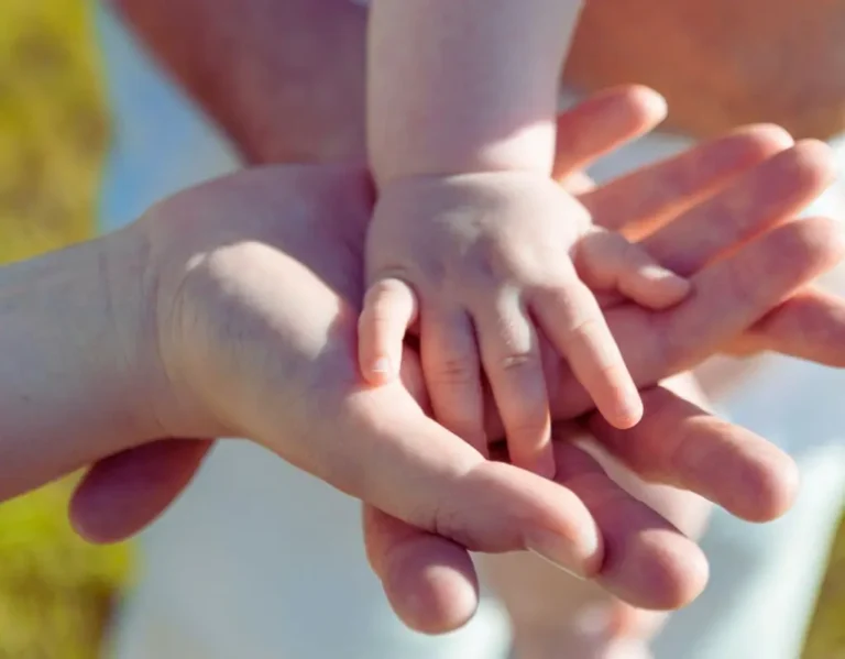 Na zdjęciu widoczne są trzy dłonie – dorosłego mężczyzny, kobiety i dziecka – ułożone jedna na drugiej w geście bliskości i zaufania. Mała rączka dziecka spoczywa na dłoniach rodziców, symbolizując opiekę, miłość i poczucie bezpieczeństwa. To obraz pełen ciepła, oddający emocjonalną więź i dobre relacje z dzieckiem. Ujęcie emanuje spokojem, delikatnością i wzajemnym wsparciem – pokazuje, jak ważna jest obecność i bliskość w budowaniu rodzinnych relacji.