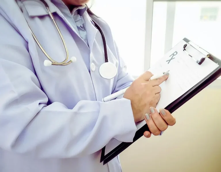 Na obrazku widoczna jest osoba w lekarskim kitlu, trzymająca w dłoni długopis i wypisująca coś na kartce przypiętej do czarnego klipbordu. Na szyi ma zawieszony stetoskop, co jednoznacznie wskazuje na pracownika ochrony zdrowia. Kartka, którą uzupełnia, przypomina formularz medyczny lub receptę – w górnej części widać symbol „Rx”. Zdjęcie budzi skojarzenia z profesjonalizmem, zaufaniem i skupieniem na pacjencie. Może ilustrować proces wystawiania dokumentów związanych z orzeczeniem o niepełnosprawności lub chorobą przewlekłą. Idealnie pasuje jako ilustracja tematu lista chorób do bezterminowego orzeczenia – podkreśla powagę i odpowiedzialność decyzji medycznych oraz ich wpływ na życie pacjenta.