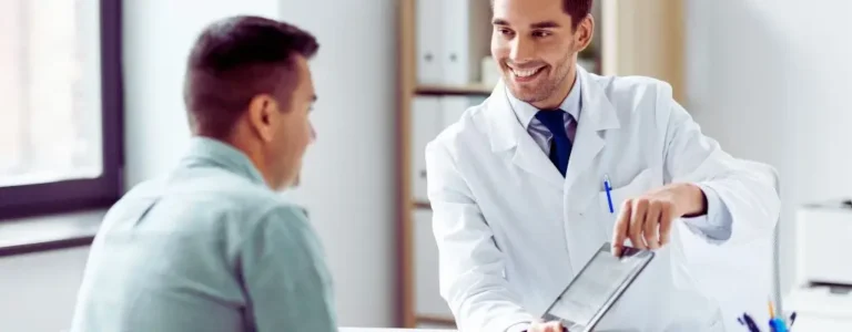 Lekarz z uśmiechem pokazuje pacjentowi informacje na tablecie w jasnym gabinecie. Obraz ilustruje profesjonalną pomoc i wsparcie w zakresie zdrowia, nawiązując do tematu: wykaz sprzętu rehabilitacyjnego refundowanego przez PFRON.