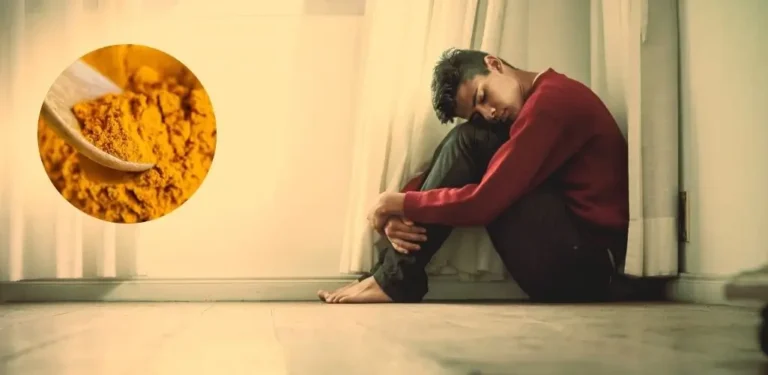 Na obrazku widzimy młodego mężczyznę siedzącego samotnie na podłodze pod oknem, z głową opartą o kolana. Ma smutny, zamyślony wyraz twarzy, co sugeruje stan przygnębienia, bezsilności lub depresji. Całość utrzymana jest w ciepłej, przytłumionej tonacji, potęgującej uczucie izolacji i bezradności. W lewym górnym rogu znajduje się okrągłe wstawienie ze zdjęciem kurkumy w proszku na drewnianej łyżce, co może sugerować powiązanie między naturalnym składnikiem a emocjonalnym stanem osoby. Obrazek ilustruje pytanie: czy kurkumina pomaga w depresji, łącząc ludzkie doświadczenie z potencjalnym naturalnym wsparciem.