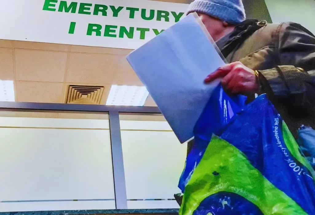 Osoba w zimowej kurtce i czapce stoi pod tablicą „EMERYTURY I RENTY”, trzymając reklamówkę i dokumenty. Kadr uchwyca codzienność osób ubiegających się o wsparcie – takie jak najczęściej przyznawane renty. Widać skupienie, może napięcie, typowe dla wizyt w urzędzie.