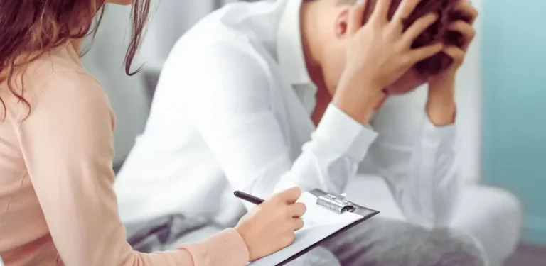 Na zdjęciu widać scenę rozmowy terapeutycznej – na pierwszym planie znajduje się kobieta z notesem i długopisem, prawdopodobnie psycholog lub psychiatra, notująca informacje. Na drugim planie mężczyzna w białej koszuli siedzi pochylony, trzyma głowę w dłoniach w geście rozpaczy lub bezradności. Jego postawa i mimika sugerują silny stres emocjonalny, przygnębienie lub objawy depresji. Obraz oddaje atmosferę intymności, wsparcia i poszukiwania pomocy w trudnym momencie życia. Idealny do zilustrowania tematu: czy na depresję można dostać orzeczenie o niepełnosprawności.