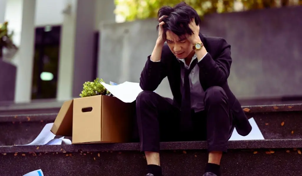 Zmartwiony mężczyzna siedzący na schodach po utracie pracy, obok kartonu z rzeczami z biura – wizualizacja tematu „jak sobie radzić z bezrobociem”.